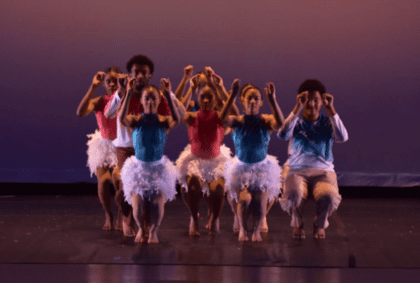 A group of young dancers on stage, joyfully raising their arms in unison, showcasing their enthusiasm and talent.